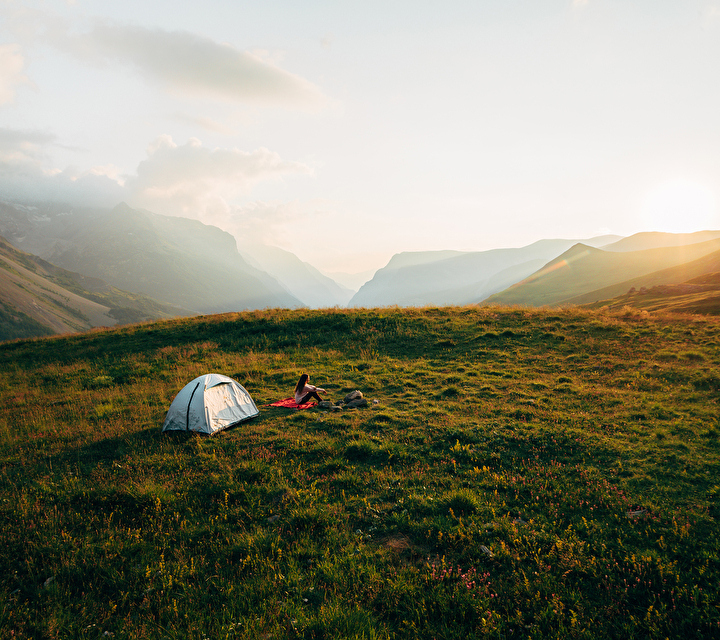 Femme assise devant sa tente posée en prairie regardant les montagnes et le coucher de soleil