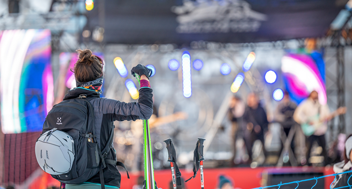 Skieuse de dos avec un sac à dos et des bâtons de ski, observant un concert en plein air sur une scène illuminée, dans une station de ski enneigée. Ambiance festive mêlant sport d'hiver et musique live.