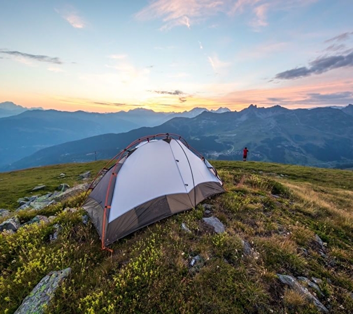 Tente plantée sur une prairie et personne observant le coucher de soleil sur les montagnes environnantes