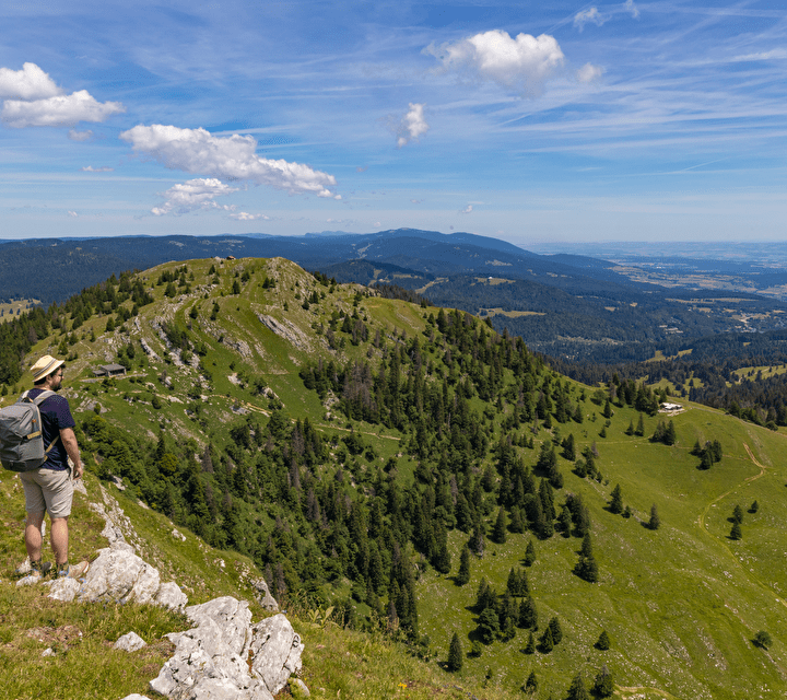 Randonneur contemplant un panorama sur les montagnes du Jura, avec des crêtes verdoyantes, un ciel bleu parsemé de nuages et des vallées s'étendant à l'horizon.