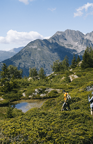 Randonnée en pleine nature dans les Alpes du Nord, avec deux marcheurs traversant des sentiers verdoyants entourés de montagnes majestueuses et d'un petit étang alpin sous un ciel bleu.