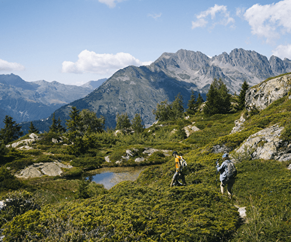 Randonnée en pleine nature dans les Alpes du Nord, avec deux marcheurs traversant des sentiers verdoyants entourés de montagnes majestueuses et d'un petit étang alpin sous un ciel bleu.