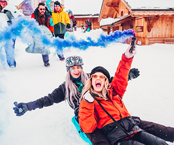 Un groupe d'amis profitant d'une journée enneigée dans une station de ski, avec des personnes en luge, une ambiance festive, et des fumigènes bleus. En arrière-plan, des chalets en bois traditionnels et une atmosphère hivernale animée.
