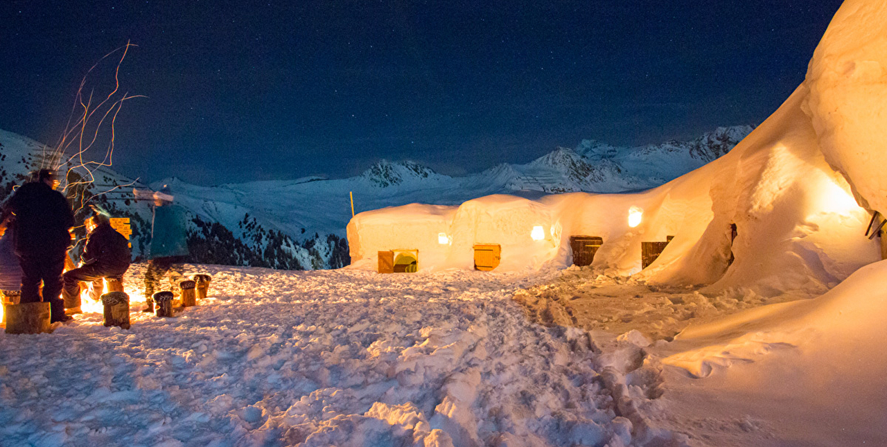 Village igloo illuminé en pleine montagne où plusieurs personnes sont réunies autour d'un feu