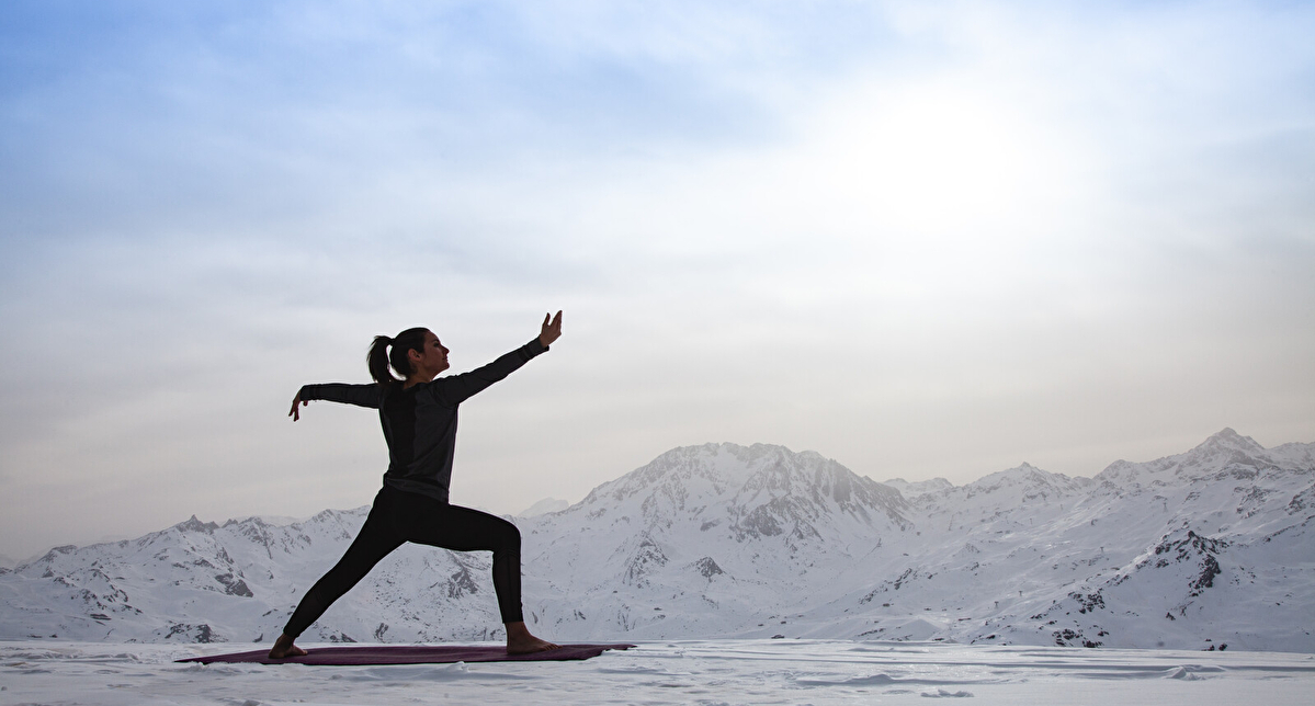 Femme pratiquant le yoga en altitude sur un tapis posé dans la neige, avec une vue panoramique sur des montagnes enneigées sous un ciel clair. Activité bien-être en pleine nature dans une station de ski.