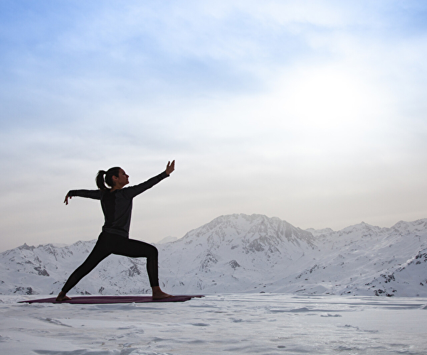 Femme pratiquant le yoga en altitude sur un tapis posé dans la neige, avec une vue panoramique sur des montagnes enneigées sous un ciel clair. Activité bien-être en pleine nature dans une station de ski.