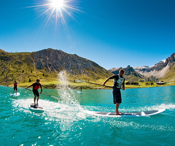 Personnes sur un lac turquoise faisant du stand-up paddle face aux montagnes verdoyantes ensoleillées