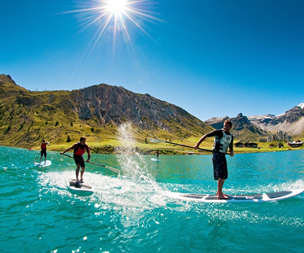 Personnes sur un lac turquoise faisant du stand-up paddle face aux montagnes verdoyantes ensoleillées