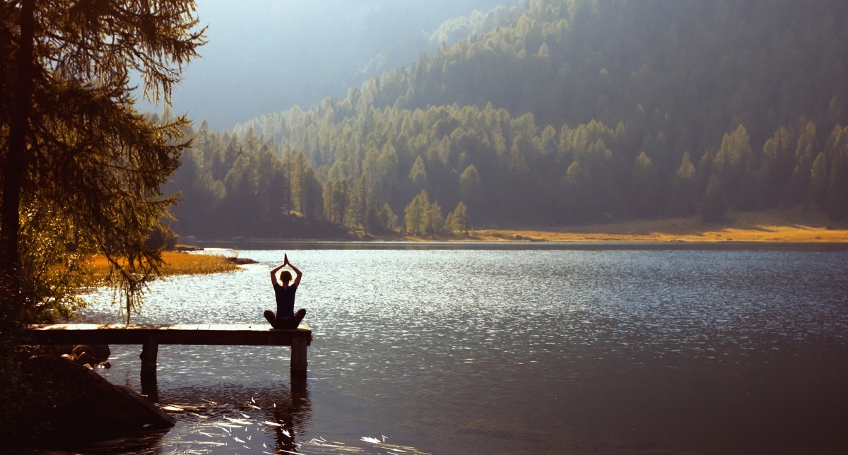 Personne en position assiste sur un ponton près d'un lac effectuant une séance de yoga face à la nature et aux montagnes