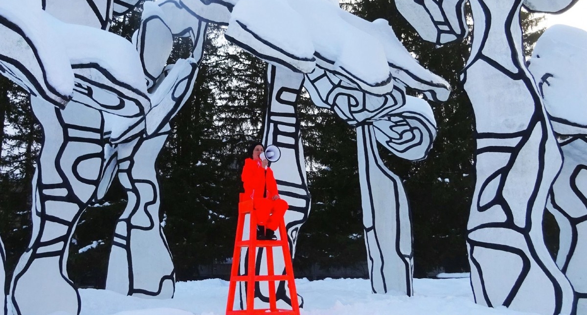 Oeuvre d'art exposée dans la neige où une jeune femme assise sur un fauteuil haut rouge utilise un mégaphone pour se faire entendre