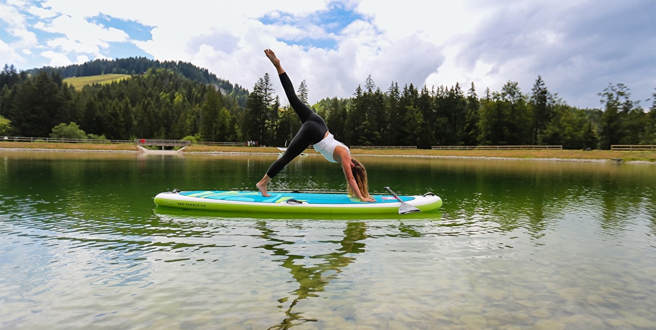 Personne effectuant une posture de yoga sur un paddle au milieu d'un lac entouré de verdure l'été