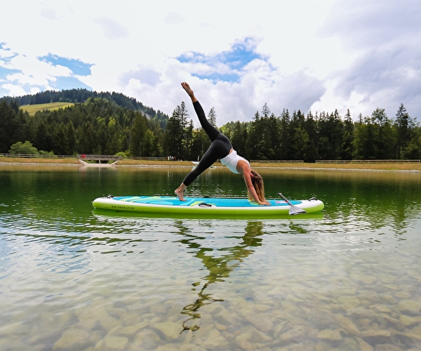 Personne effectuant une posture de yoga sur un paddle au milieu d'un lac entouré de verdure l'été