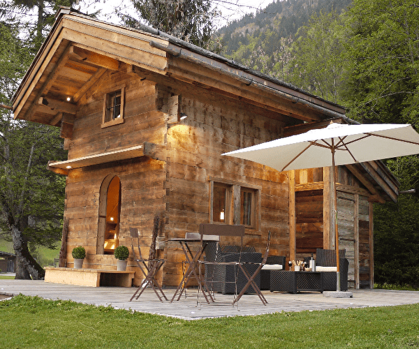 Vue extérieure sur un chalet en bois et son espace terrasse avec canapé, fauteuils et parasol en pleine nature