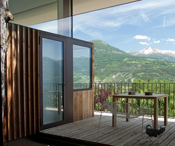 Vue sur un chalet d'habitation et sa terrasse extérieure où s'entreposent une table et un fauteuil face à un paysage de montagne