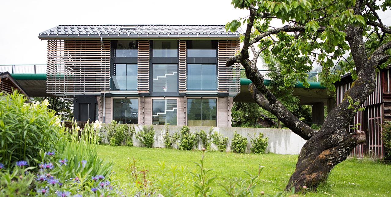 Vue sur une habitation moderne avec bardage bois extérieur et larges fenêtres donnant sur un jardin arboré et fleuri