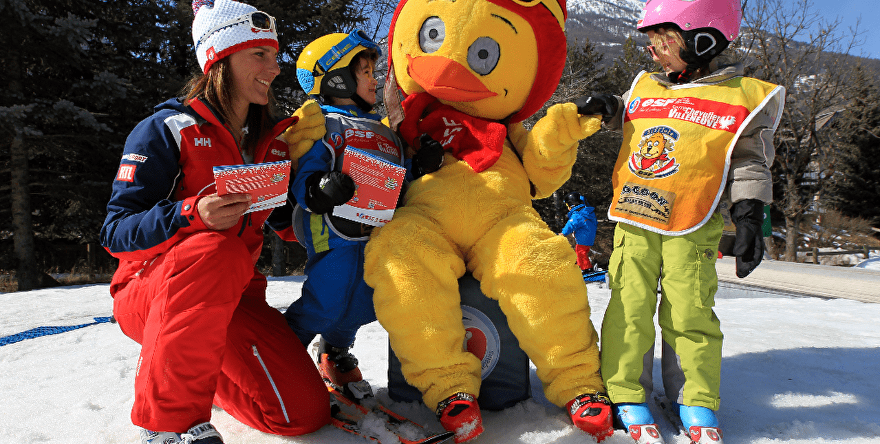 Groupe d'enfants autour d'une mascotte canard avec leur monitrice de ski sur une piste de ski l'hiver