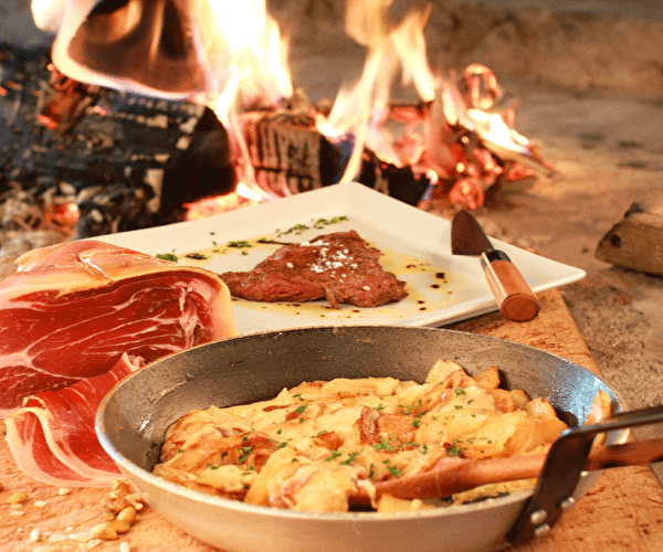 Présentation d'un plat réalisé à partir de viande, de jambon fumé et de pommes de terre gratinées autour d'un feu de bois