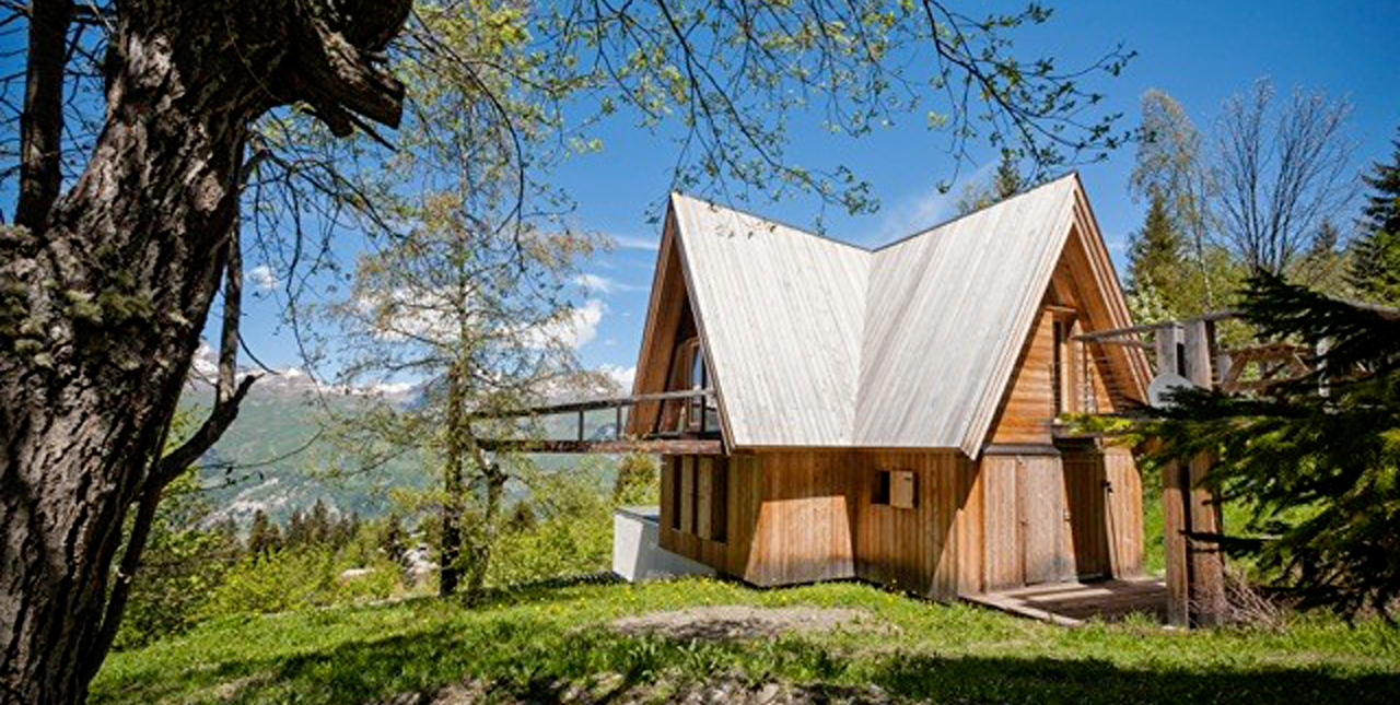 Vue sur un chalet d'habitation en bois avec balcons dans un jardin entouré d'arbres face à la montagne