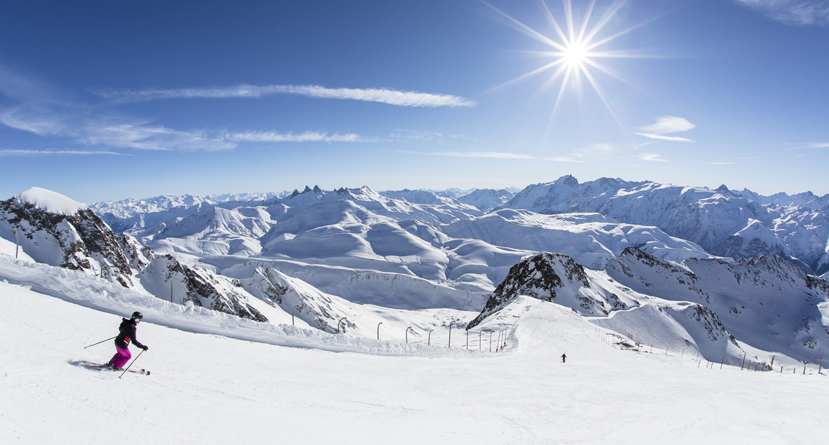 Skieurs descendant une piste de ski devant un panorama époustouflant fait de pics et de montagnes enneigées sous le soleil