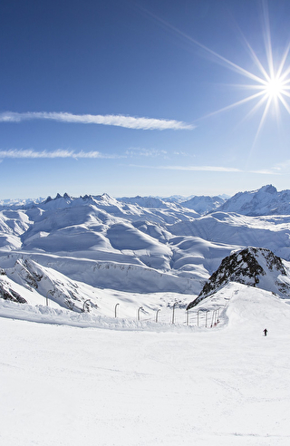 Skieurs descendant une piste de ski devant un panorama époustouflant fait de pics et de montagnes enneigées sous le soleil