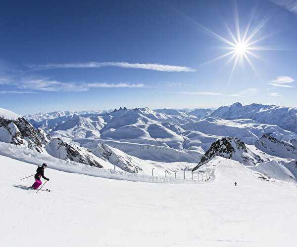Skieurs descendant une piste de ski devant un panorama époustouflant fait de pics et de montagnes enneigées sous le soleil