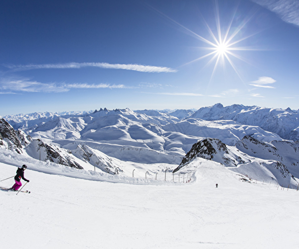 Skieurs descendant une piste de ski devant un panorama époustouflant fait de pics et de montagnes enneigées sous le soleil
