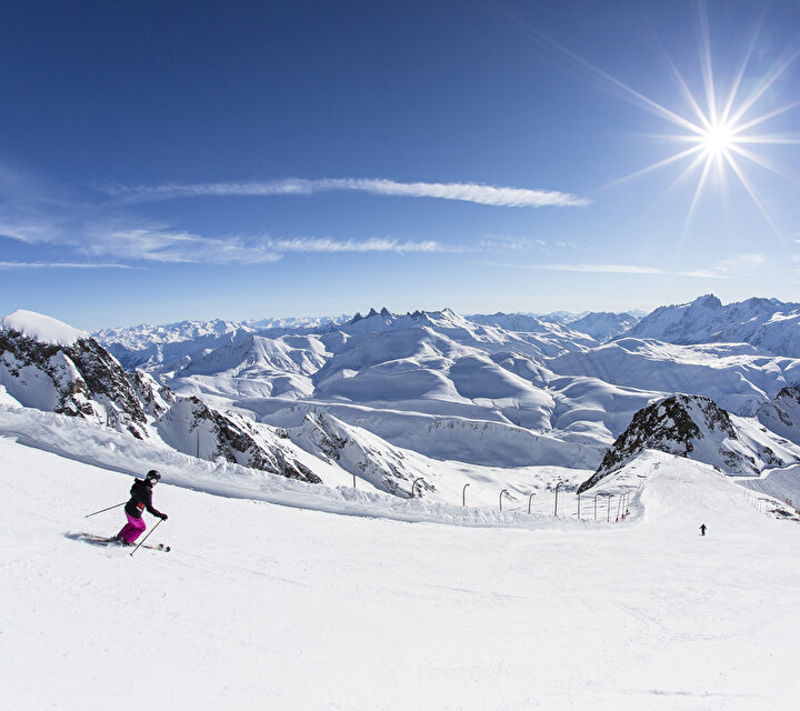 Skieurs descendant une piste de ski devant un panorama époustouflant fait de pics et de montagnes enneigées sous le soleil