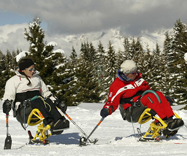 deux personnes assises dans des fauteuils handi-ski descendant une piste de montagne enneigée proche de sapins