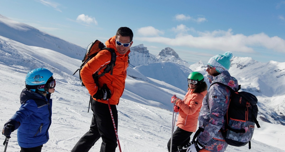 Adultes et enfants équipés de tenues pour skier, de casques, lunettes et sac à dos sur une piste face aux montagnes enneigées