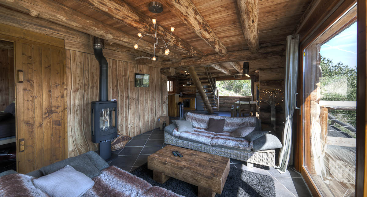 Vue intérieure d'un chalet en bois avec espace détente, salon, pôele chauffant et baie vitrée donnant sur l'extérieur