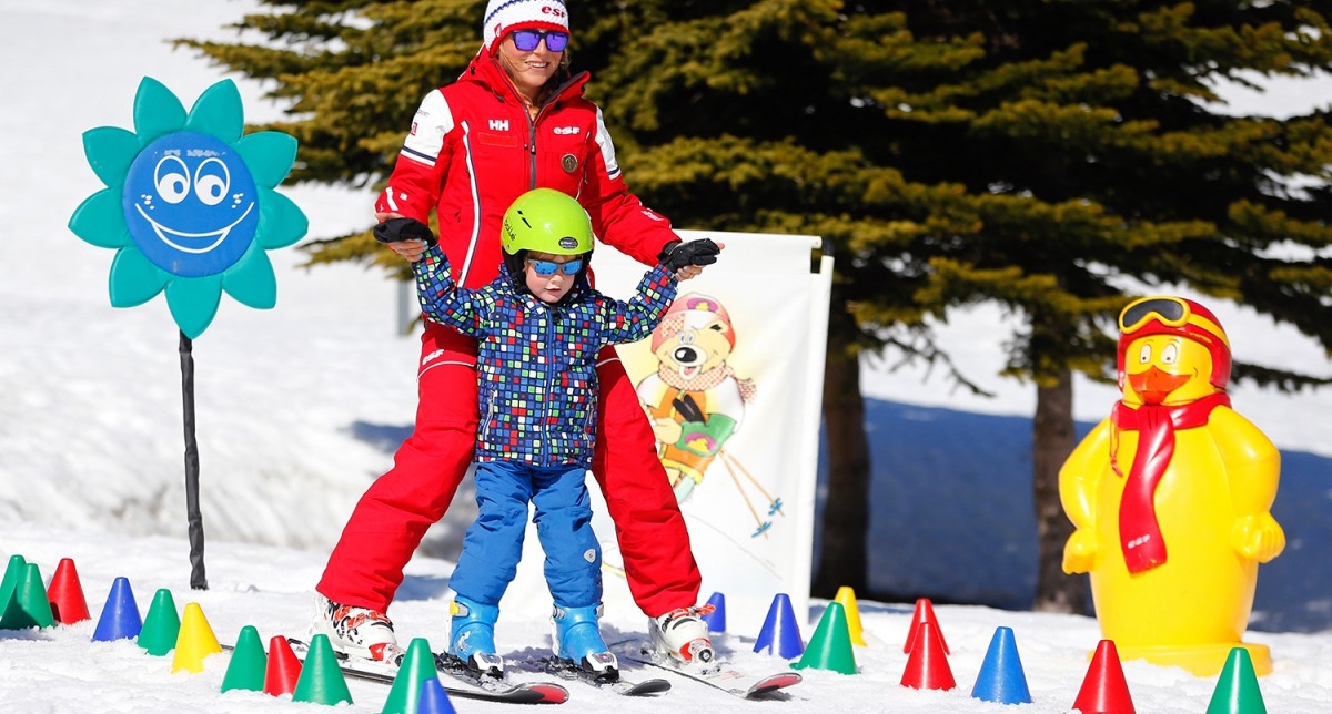 Monitrice de ski ESF apprenant à un enfant à descendre une piste entre des plots de couleurs sur un espace dédié aux jeunes