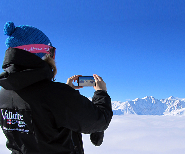 Personne habillée chaudement et portant un masque de ski réalisant des photos de la montagne et des sommets enneigés sous un ciel bleu