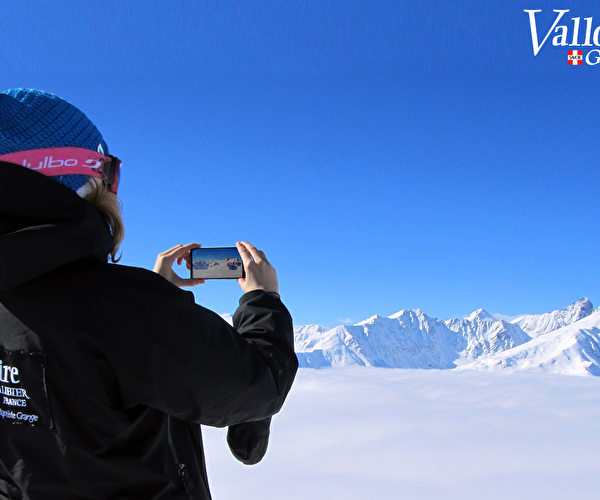 Personne habillée chaudement et portant un masque de ski réalisant des photos de la montagne et des sommets enneigés sous un ciel bleu