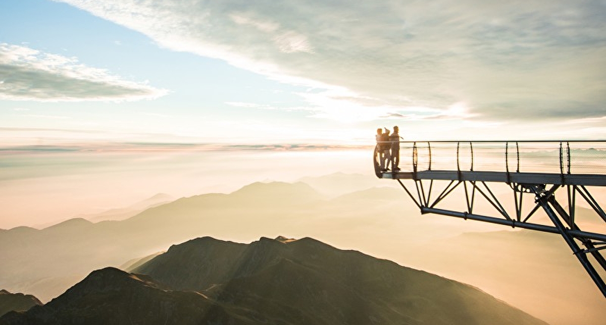 Personnes postées au bout d'une passerelle métallique offrant une vue panoramique sur les montagnes au coucher du soleil