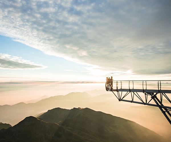 Personnes postées au bout d'une passerelle métallique offrant une vue panoramique sur les montagnes au coucher du soleil