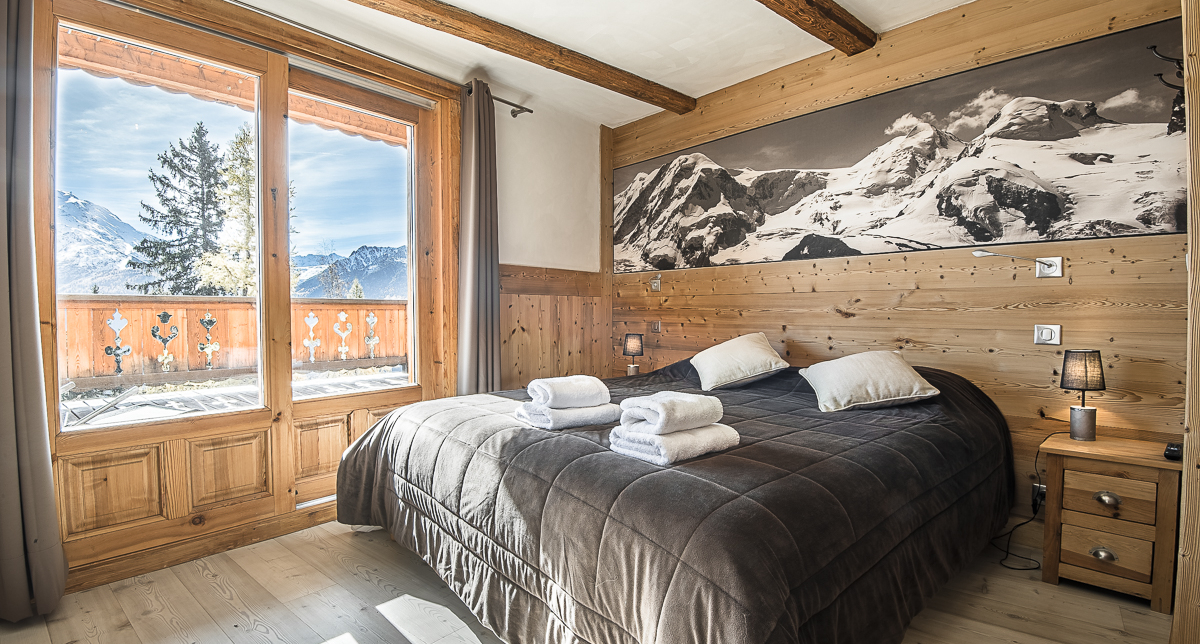 Vue intérieure d'une chambre à coucher avec lit double dont la baie vitrée donne sur un balcon face aux montagnes enneigées