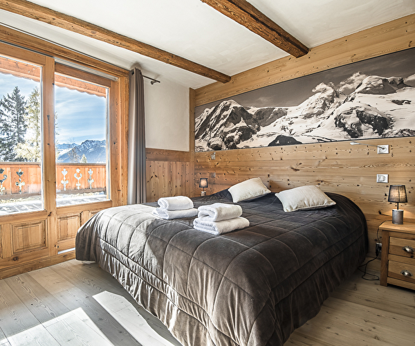 Vue intérieure d'une chambre à coucher avec lit double dont la baie vitrée donne sur un balcon face aux montagnes enneigées