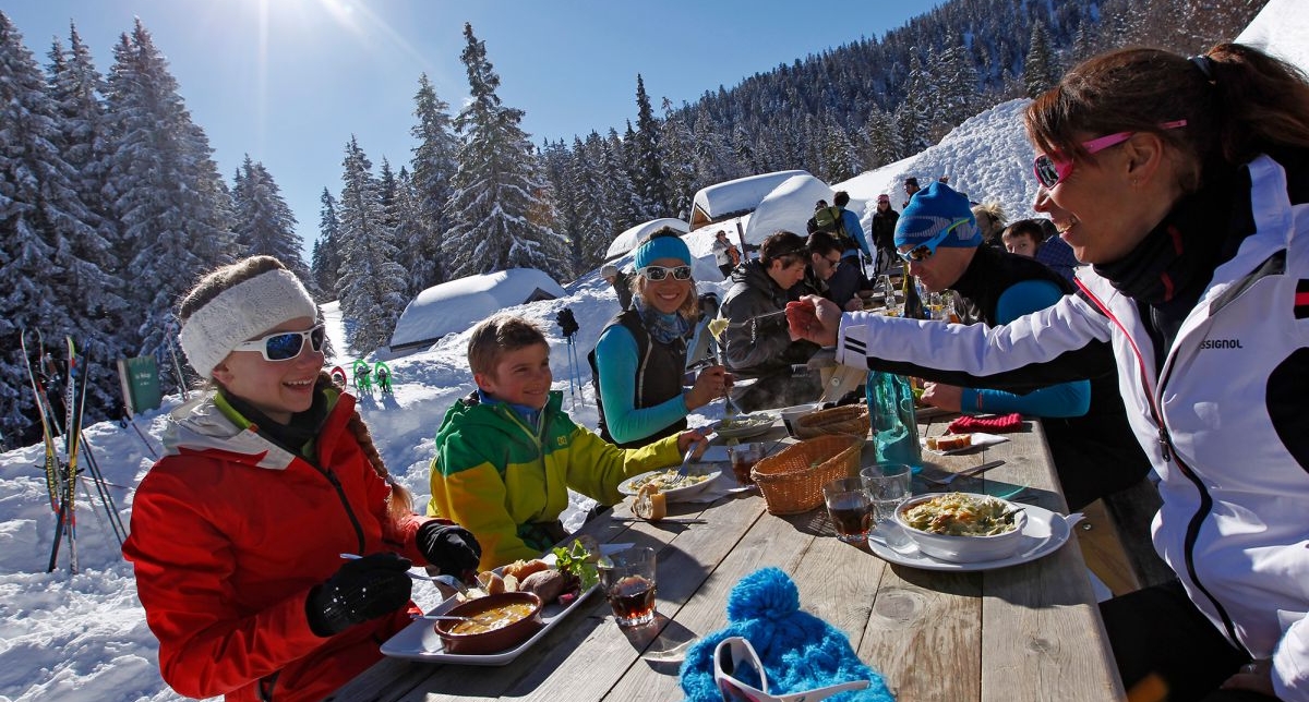 Grande tablée familiale en extérieur où adultes et enfants dégustent des spécialités de montagne sous le soleil