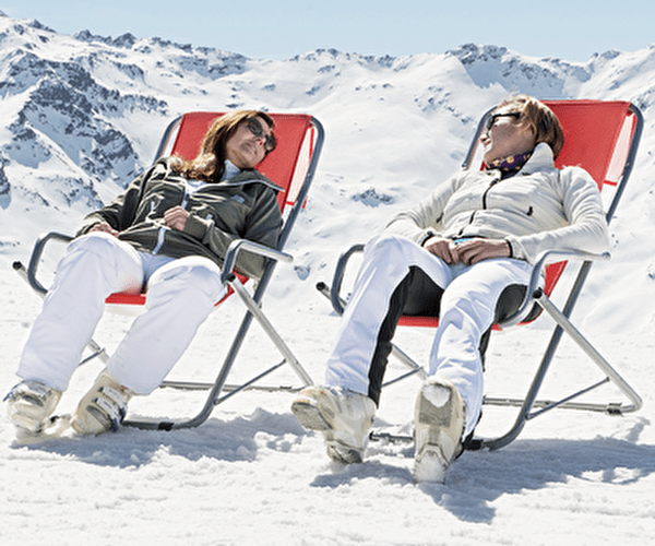 Personnes en tenues de ski assises dans des transats oranges sur la neige face aux montagnes enneigées et sous le soleil de printemps